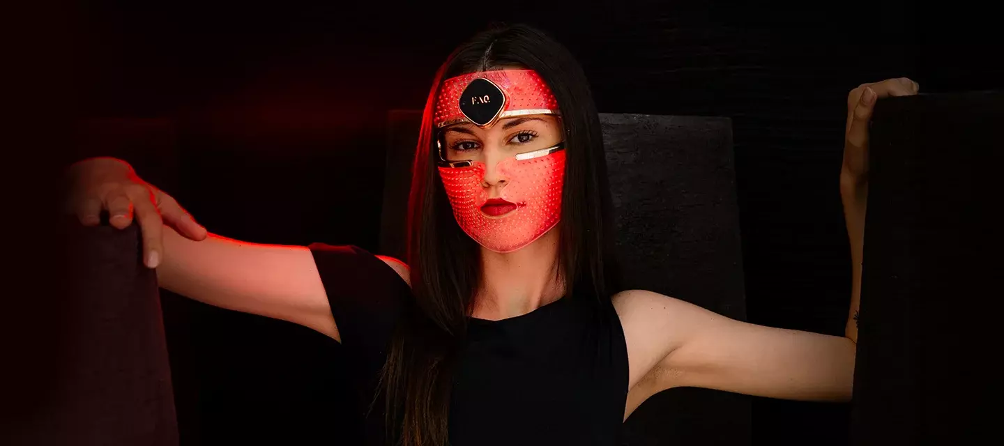 A young woman with dark hair and eyes in a sleveless black dress on black background wearing a red LED face mask 